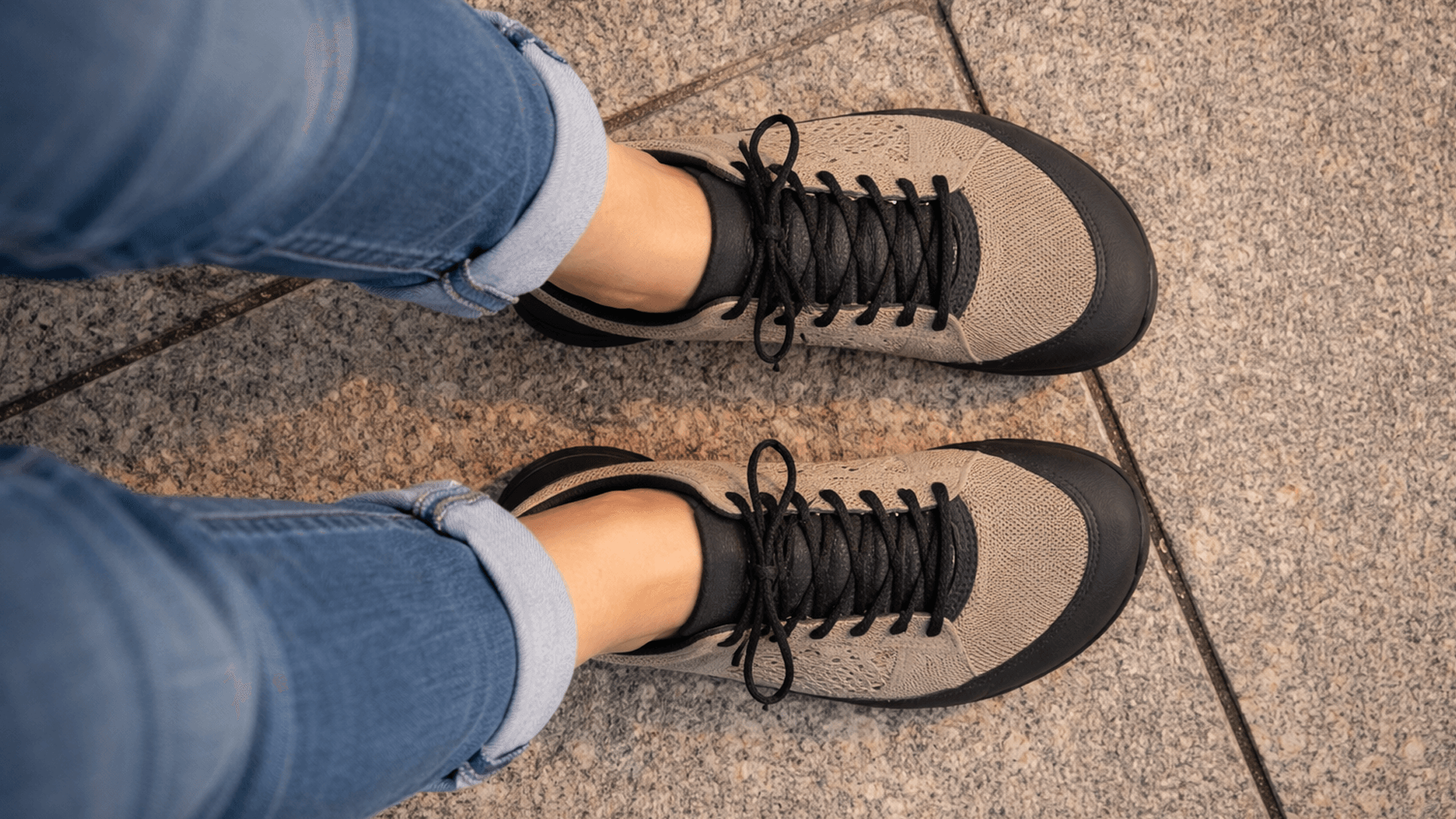 Vista superior de una persona usando calzado barefoot con puntera amplia y suela plana, de pie sobre una vereda urbana de baldosas.